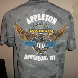 Może przedstawiać: Szara koszulka z logo Harley Davidson i tekstem "Appleton, WI". Logo przedstawia orła z rozpostartymi skrzydłami, słowa "Harley Davidson" i tekst "The Great American Freedom Machine".