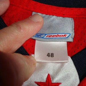 May include: Red and black Reebok jersey with a white star on the front. The jersey has a size tag that says "48" and "Reebok".