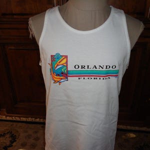 May include: White tank top with a colorful graphic featuring two dolphins and the text "ORLANDO FLORIDA". The design includes orange, yellow, blue, and green elements, with a red and blue stripe below the city name.