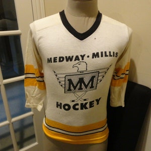 May include: White hockey jersey with black and yellow stripes. The jersey has a black eagle logo with the letters "MM" in the center and the text "MEDWAY MILLIS HOCKEY" printed on the front.