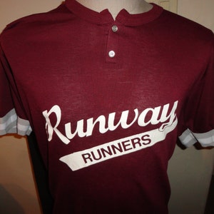 Puede incluir: Camiseta color burdeos con mangas blancas y un gráfico blanco que dice "Runaway Runners" en una fuente retro.