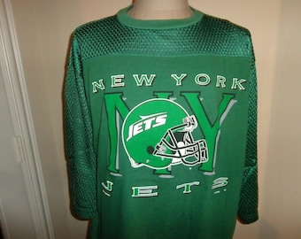 以下が含まれることがあります： 白文字で「NEW YORK JETS」と書かれた緑色の長袖シャツ。シャツには、中に「JETS」という文字が入った緑色のフットボールヘルメットのグラフィックが描かれています。袖と背中上部はメッシュ素材です。