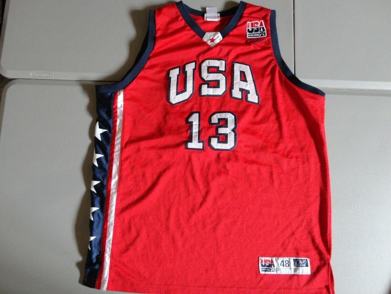 May include: Red basketball jersey with white "USA" and "13" lettering, blue and white star accents on the sides, and a small "USA" tag on the bottom.