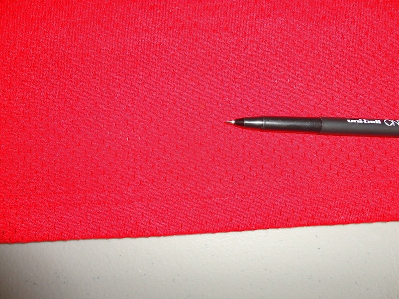 May include: Close-up of a red mesh fabric with a small pen lying on top of it. The pen is black with white lettering that says "uni-ball ON".