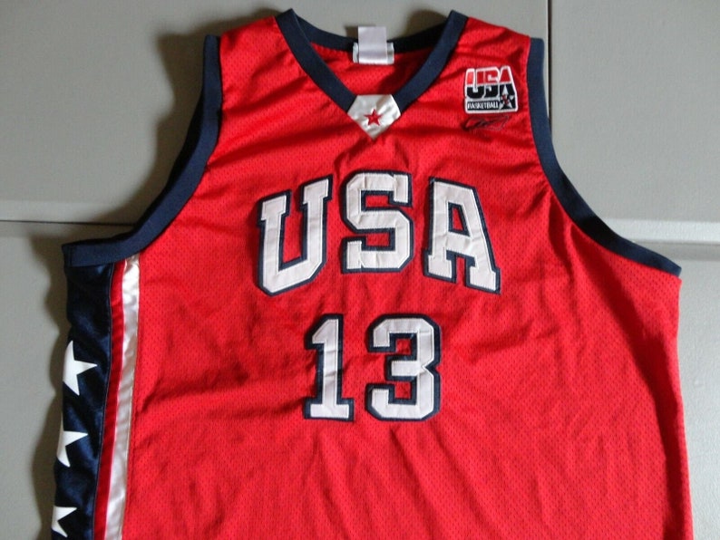 May include: Red basketball jersey with white "USA" and "13" lettering, blue trim, and a small USA Basketball logo on the right shoulder.