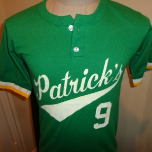 Può includere: Maglia da baseball verde a maniche corte con bordini bianchi e gialli. La maglia ha un "Patrick's" bianco con una linea curva bianca e il numero "9" in bianco.