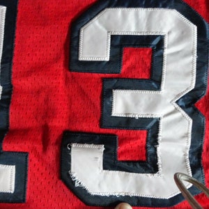 May include: Close-up of a red jersey with the number "13" sewn on in white with a navy blue outline.