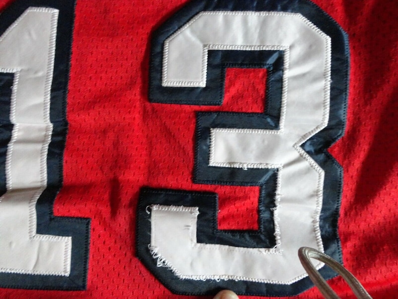 May include: Close-up of a red jersey with the number "13" sewn on in white with a navy blue outline.