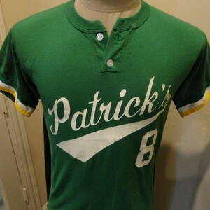 Può includere: Maglia da baseball verde con bordini bianchi e gialli. La maglia ha il testo "Patrick's" in lettere bianche corsive con un numero 8 bianco sul davanti.