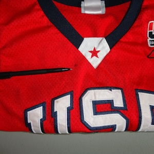May include: Red basketball jersey with white and blue trim. The jersey has the number "15" on the front and the words "USA Basketball" on the right shoulder.