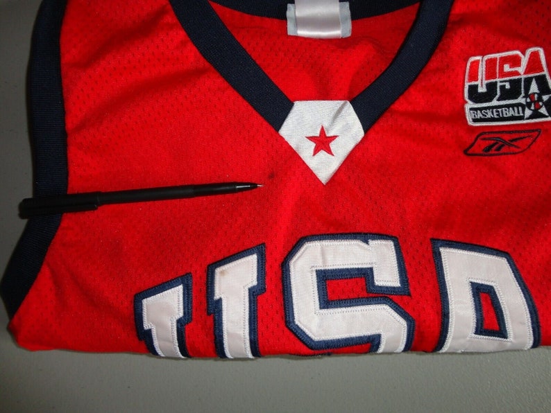 May include: Red basketball jersey with white and blue trim. The jersey has the number "15" on the front and the words "USA Basketball" on the right shoulder.