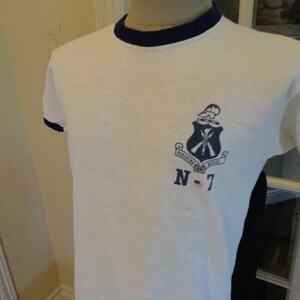 May include: White t-shirt with a blue ringer neck and a blue and white crest with the text "PERSHING RIFLES" and "N-7".