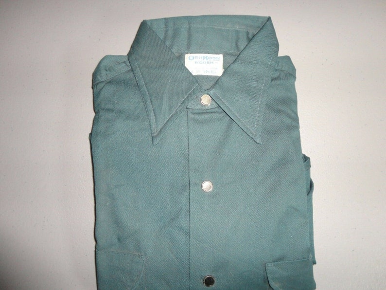 May include: A dark green long-sleeved shirt with a button-down collar and a single pocket on the left side. The shirt has a label that reads "Orion Made in Korea".