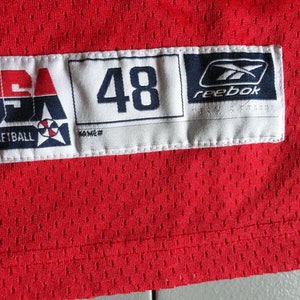 May include: A white tag with the text "USA Basketball" and a basketball logo. The tag also has the number "48" and the Reebok logo. The tag is attached to a red jersey.
