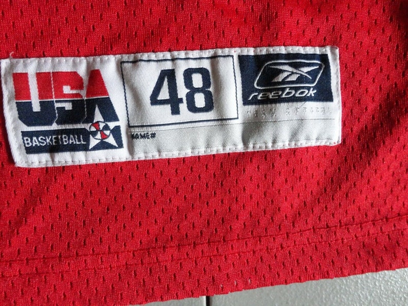 May include: A white tag with the text "USA Basketball" and a basketball logo. The tag also has the number "48" and the Reebok logo. The tag is attached to a red jersey.
