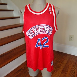 May include: Red basketball jersey with blue and white lettering that says "SIXERS 42" and a white Champion logo on the front.