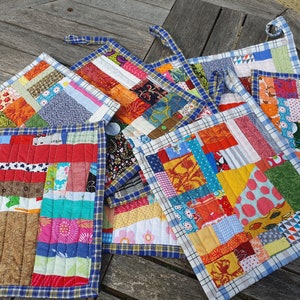Op de afbeelding: Zes kleurrijke patchwork ovenwanten met een blauwe en witte geruite rand. Elke ovenwant is gemaakt van verschillende stoffen en patronen.