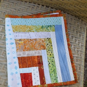 Könnte beinhalten: Eine Patchwork-Decke mit einer Vielzahl von bunten Stoffen, darunter Blau, Grün, Orange und Weiß. Die Decke besteht aus Quadraten und Rechtecken in verschiedenen Größen und Mustern. Die Decke liegt auf einem Korbstuhl.