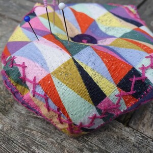 May include: A colorful fabric pincushion with a geometric pattern in shades of pink, orange, yellow, blue, green, and black. The pincushion has a pink border and is filled with pins with colorful plastic heads.