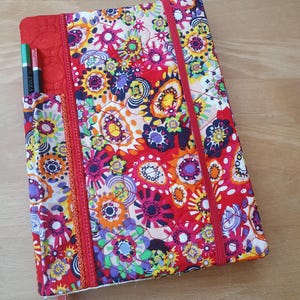 Könnte beinhalten: Ein gestepptes Notizbuch mit einem lebendigen Blumenmuster in Rot, Rosa, Gelb und Blau. Das Notizbuch hat einen roten Gummibandverschluss und eine rote Tasche mit zwei Stiften. Ein rosa Lesezeichen ist sichtbar.