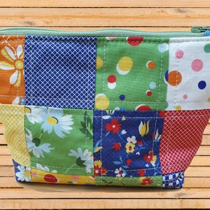 Könnte beinhalten: Eine gepatchte Stofftasche mit Reißverschluss und grünem Zipper. Die Tasche besteht aus verschiedenen Stoffen mit Blumen-, Punkte- und geometrischen Mustern.