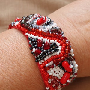 Può includere: Un braccialetto rosso, bianco e grigio con un motivo geometrico. Il braccialetto è realizzato con piccole perline rotonde e ha un design unico e moderno.