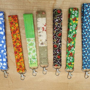 Może przedstawiać: Siedem kolorowych breloków z tkaniny ze srebrnymi kółkami. Breloki mają różne wzory kwiatowe w kolorze niebieskim, zielonym, pomarańczowym, czerwonym i czarnym.