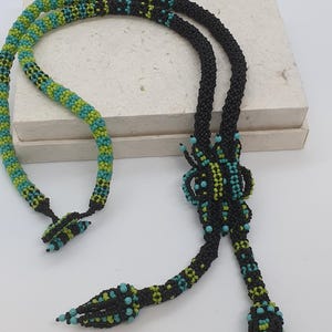 Könnte beinhalten: Eine lange Lariat-Halskette aus kleinen schwarzen, lindgrünen und türkisfarbenen Saatperlen. Das Design zeigt ein zentrales Knotenelement mit zwei herabhängenden Enden, die jeweils in einer spitz zulaufenden, perlenbesetzten Quaste enden.