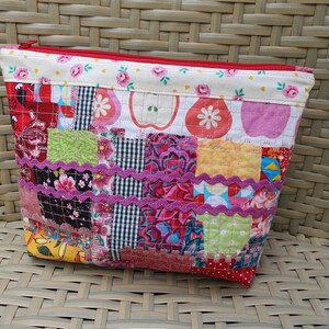 Peut inclure: Une pochette zippée en patchwork avec une fermeture éclair rouge et un imprimé floral et de pommes. La pochette est composée de différents carrés de tissu de couleurs et de motifs variés, avec une bordure décorative violette.