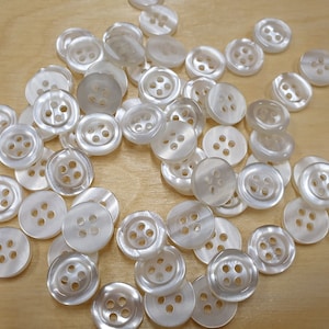 Può includere: Un grande gruppo di bottoni di plastica bianca con quattro fori ciascuno. I bottoni sono disposti in modo casuale su una superficie di legno marrone chiaro.