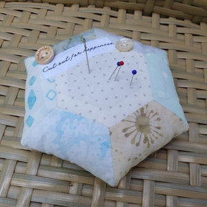Könnte beinhalten: Ein sechseckiger Nadelkissen mit weißem Hintergrund und blau-beige gemustertem Stoff. Das Nadelkissen hat ein Etikett mit der Aufschrift "Cut out for happiness" und drei hineingesteckte Nadeln.