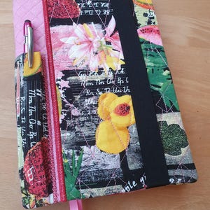 Op de afbeelding: Een gewatteerd notitieboek met een kleurrijk bloemen- en fruitpatroon. De kaft heeft een roze pennenhouder met een zilveren pen, een zwarte elastische band en een roze lint bladwijzer. Het ontwerp bevat tekst en illustraties van fruit en bloemen.