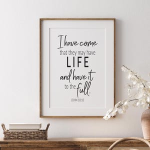 May include: A framed print with a white background and black text. The text reads "I have come that they may have LIFE and have it to the full. JOHN 10:10". The print is sitting on a wooden dresser with a wicker basket and a wooden chair.