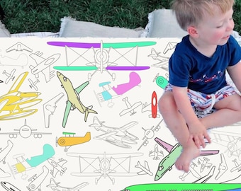 May include: A white playmat with a black outline of various airplanes. The airplanes are colored in with different colors, including green, blue, yellow, pink, and purple. The playmat is on a green grassy surface.