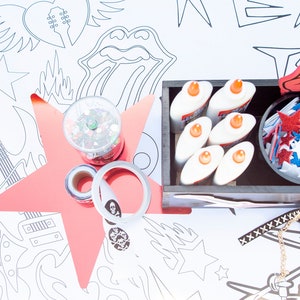 Puede incluir: Una mesa de manualidades con un mantel blanco cubierto de dibujos en blanco y negro con temática de rock and roll. Hay una gran estrella rosa, un recipiente de purpurina, un rollo de cinta adhesiva, un recipiente de pegamento en barra, un recipiente de estrellas rojas y azules en forma de estrella y un recipiente de rotuladores de colores.