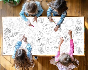 Puede incluir: Cuatro niños están sentados alrededor de un gran mantel blanco con un diseño para colorear de temática espacial. El mantel presenta planetas, estrellas, cohetes, extraterrestres y astronautas. Los niños están coloreando el diseño con marcadores.