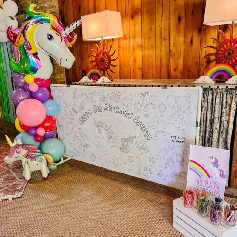 May include: A white backdrop with a unicorn theme for a 5th birthday party. The backdrop is decorated with a rainbow, stars, and the words "Happy 5th Birthday Everly". There is a table with a sign that says "Color Away!" and a variety of colored crayons in a white wooden box.