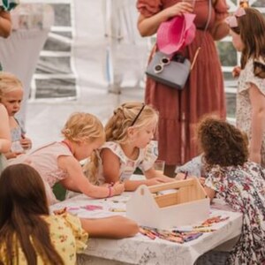 May include: A group of children are sitting at a table coloring with crayons. There is a white wooden toolbox on the table with crayons inside. The children are wearing colorful clothing and are having fun.
