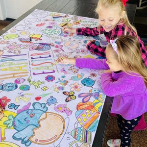 May include: Two children color a large white paper tablecloth with colorful crayons. The tablecloth features an Easter theme with the words "Happy Easter" and various Easter-themed illustrations, including eggs, bunnies, and chicks.