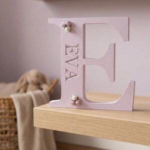 May include: A large, light purple, freestanding letter E with the name EVA engraved vertically in the centre. The letter is decorated with small, delicate clusters of pink and white artificial flowers at the top and bottom corners.