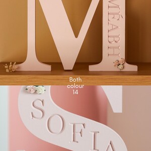 May include: A light pink wooden letter M with the name M&Eacute;ABH engraved vertically and decorated with small fabric flowers, and a light pink wooden letter S with the name SOFIA engraved diagonally and decorated with small fabric flowers.