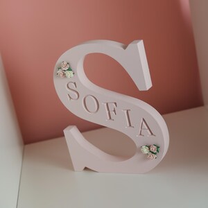 May include: A light pink, freestanding letter S featuring the name SOFIA embossed in the centre. The letter is decorated with small, delicate clusters of artificial cream and pink roses positioned at the top left and bottom right curves.