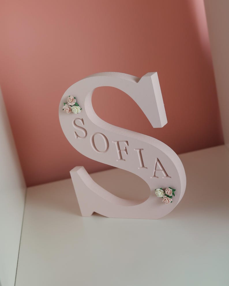 May include: A light pink, freestanding letter S featuring the name SOFIA embossed in the centre. The letter is decorated with small, delicate clusters of artificial cream and pink roses positioned at the top left and bottom right curves.