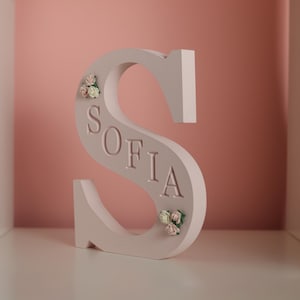 May include: A light pink, freestanding letter S featuring the name SOFIA embossed in capital letters along its centre. The letter is decorated with small, delicate clusters of artificial pink and white roses at the top left and bottom right.