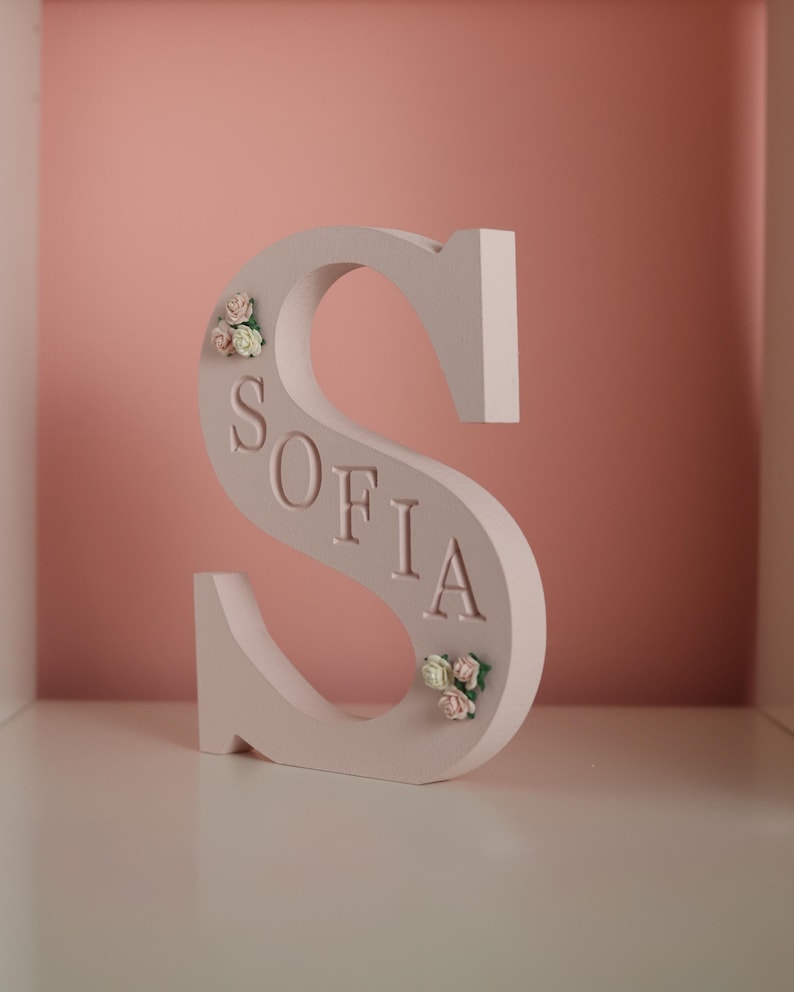 May include: A light pink, freestanding letter S featuring the name SOFIA embossed in capital letters along its centre. The letter is decorated with small, delicate clusters of artificial pink and white roses at the top left and bottom right.