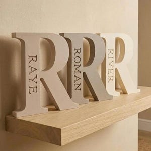 May include: Three freestanding capital letter R ornaments made of solid material in varying shades of beige and cream. Each letter features a name engraved vertically into the front surface: Raye, Roman, and River, displayed in a serif font.