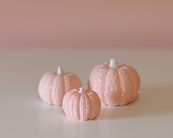 May include: Three pink pumpkins of varying sizes, with a sparkly finish, are arranged on a white surface. The pumpkins are a light shade of pink and have a slightly textured surface.