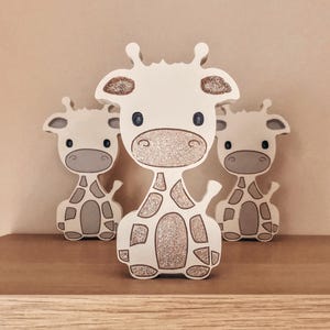 May include: Three wooden giraffe ornaments. The giraffes are white with brown spots and glitter detailing. The central giraffe is larger than the two either side. The ornaments are displayed on a brown surface against a neutral background.
