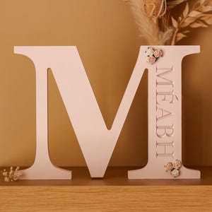 May include: A large, light pink, freestanding letter M made of wood. The right vertical bar of the letter features the name M&Eacute;ABH engraved in capital letters, accented with small, delicate fabric flowers at the top and bottom of the name.