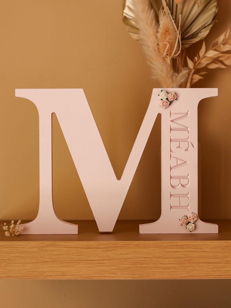 May include: A large, light pink, freestanding letter M made of wood. The right vertical bar of the letter features the name M&Eacute;ABH engraved in capital letters, accented with small, delicate fabric flowers at the top and bottom of the name.